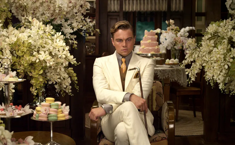 Man in wit pak zit zelfverzekerd tussen bloemen en gebak, still uit The Great Gatsby